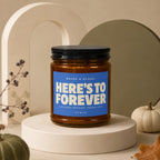 Candle with blue label reading "RAISE A GLASS: HERE'S TO FOREVER AND EVERY ORDINARY TUESDAY IN IT VOWED".