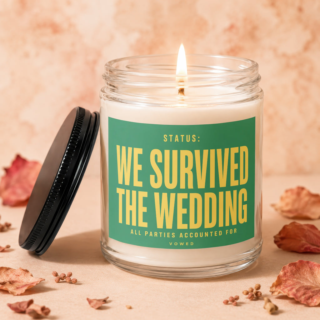 Glass candle with a lit wick and a green label reading "Status: We Survived the Wedding."