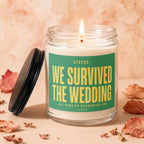 Glass candle with a lit wick and a green label reading "Status: We Survived the Wedding."