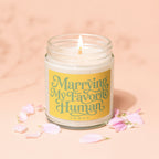 Candle with "Marrying My Favorite Human" text on yellow label, lit wick, pink petals.