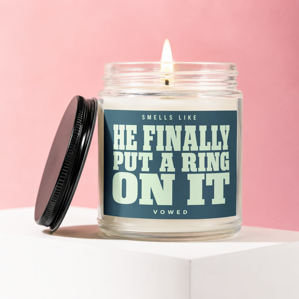 Glass candle with lit wick and black lid, text reads "Smells Like He Finally Put A Ring On It Vowed"