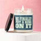 Glass candle with lit wick and black lid, text reads "Smells Like He Finally Put A Ring On It Vowed"