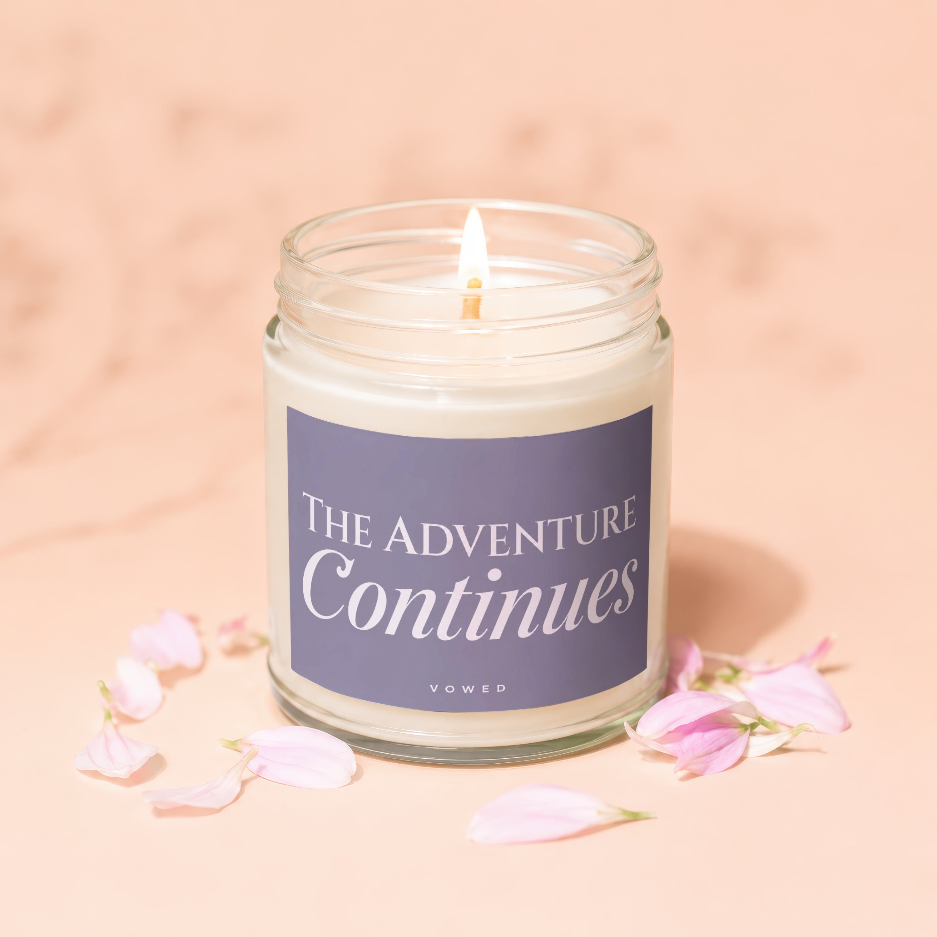 Candle with lit wick and label reading "THE ADVENTURE Continues" among flower petals.