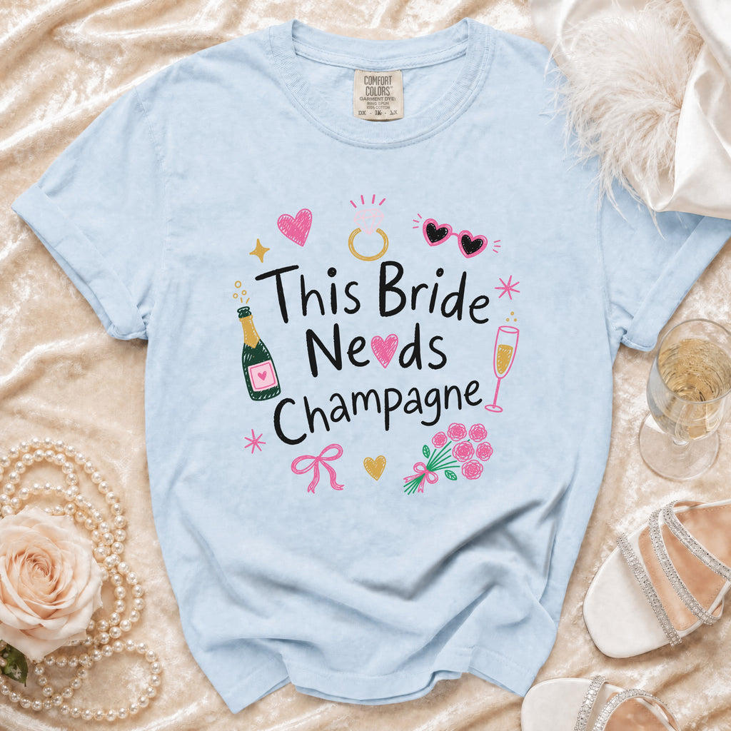 Pale blue t-shirt with "This Bride Needs Champagne" graphic and festive icons.