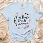 Pale blue t-shirt with "This Bride Needs Champagne" graphic and festive icons.