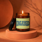Lit candle in amber jar with "Engayged | Vowed | Hand Poured" label on pedestal.