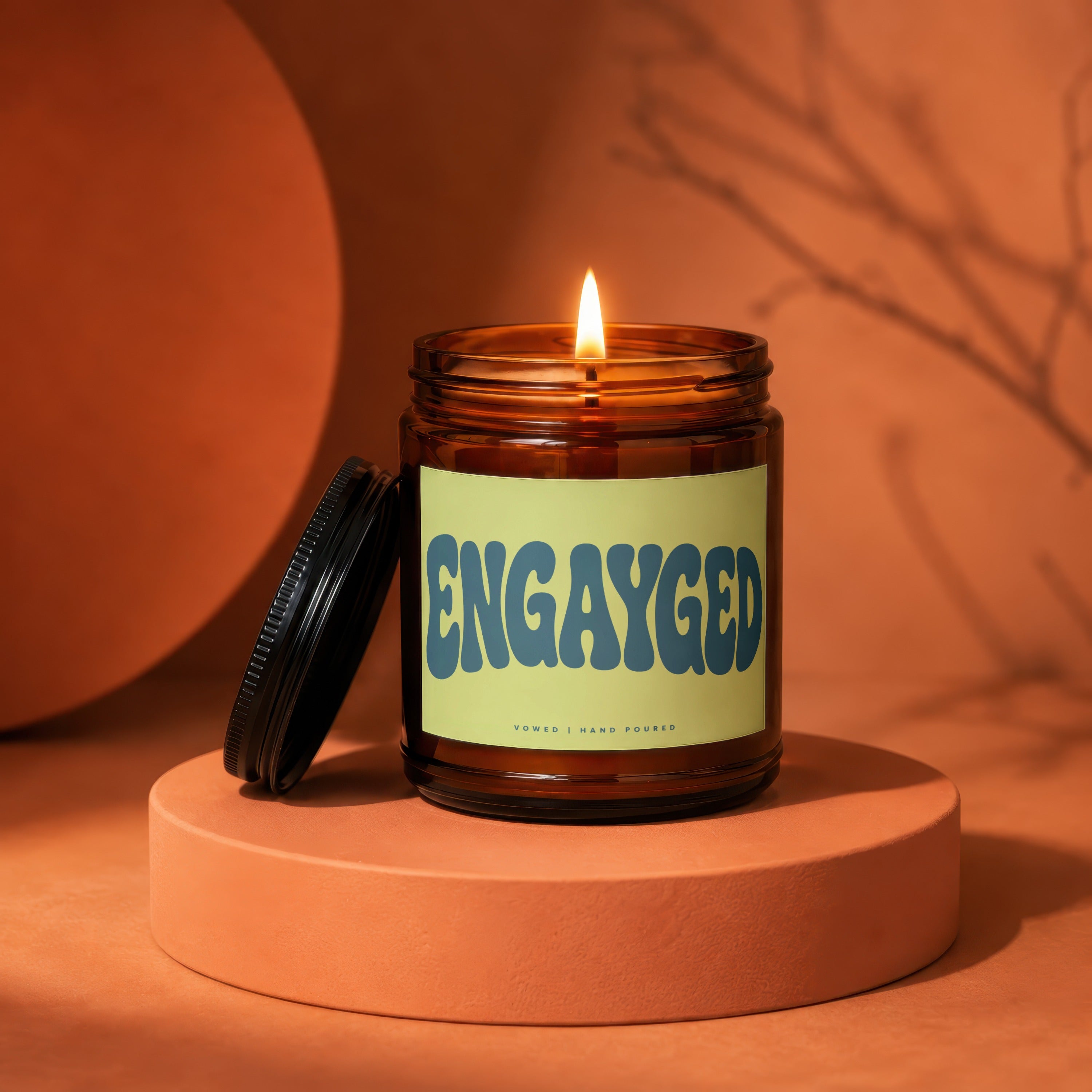 Lit candle in amber jar with "Engayged | Vowed | Hand Poured" label on pedestal.