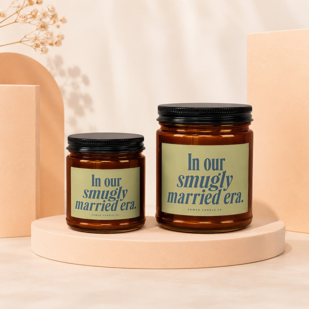 Two amber glass candles with black lids, featuring "In our smugly married era" text.