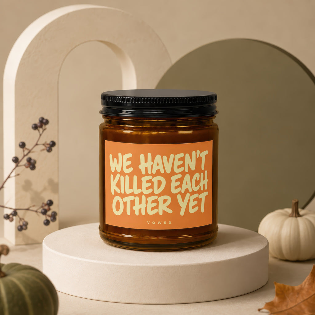 Amber jar candle with black lid displaying "WE HAVEN'T KILLED EACH OTHER YET" text.