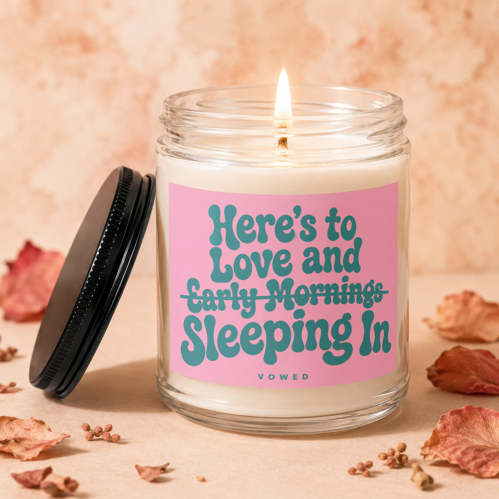 Glass candle with pink label that reads "Here's to Love and Sleeping In."