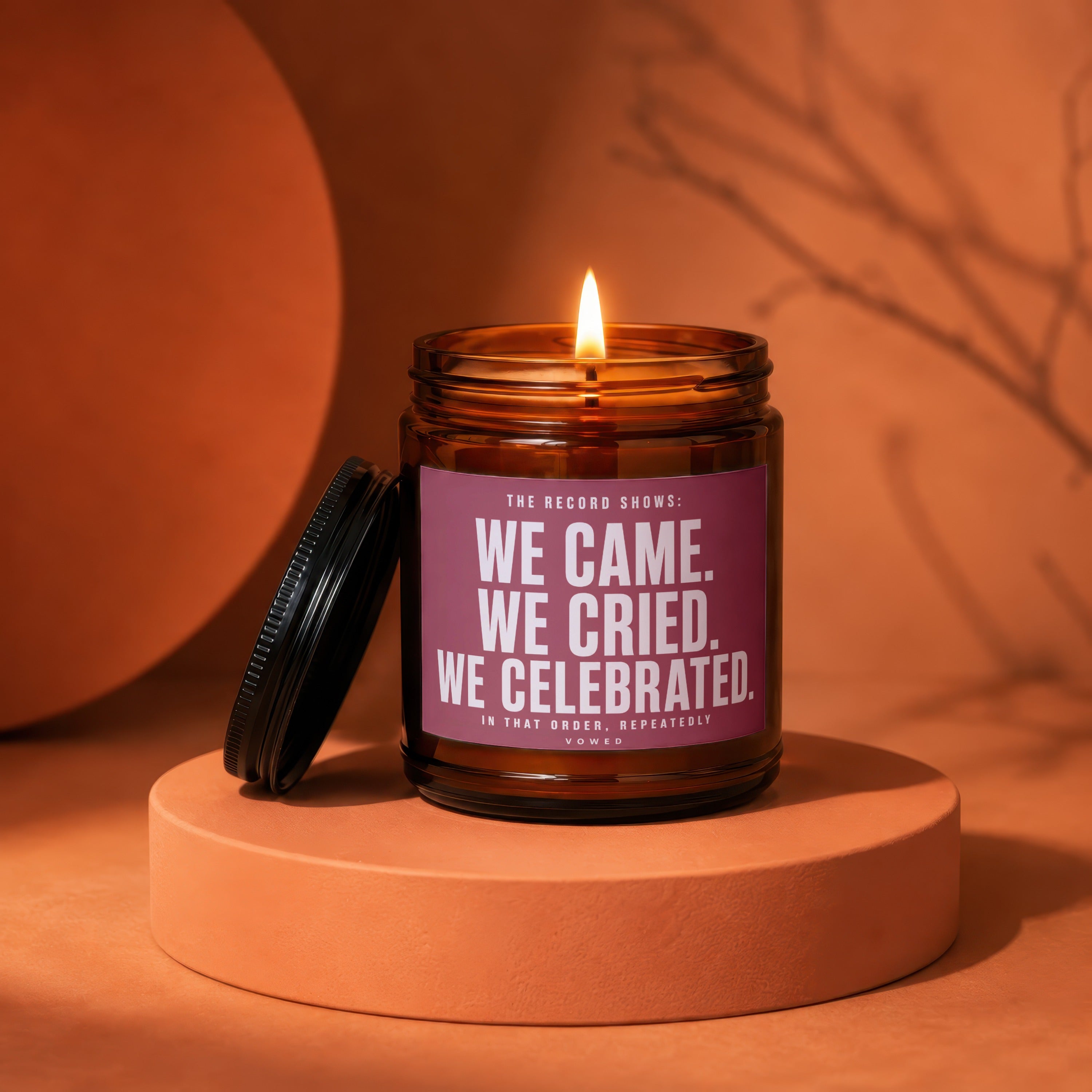Amber glass candle with lit flame and a purple label that reads "We came. We cried. We celebrated."