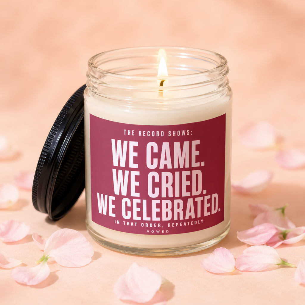 Glass candle with dark lid, lit wick, and text "WE CAME. WE CRIED. WE CELEBRATED. VOWED."