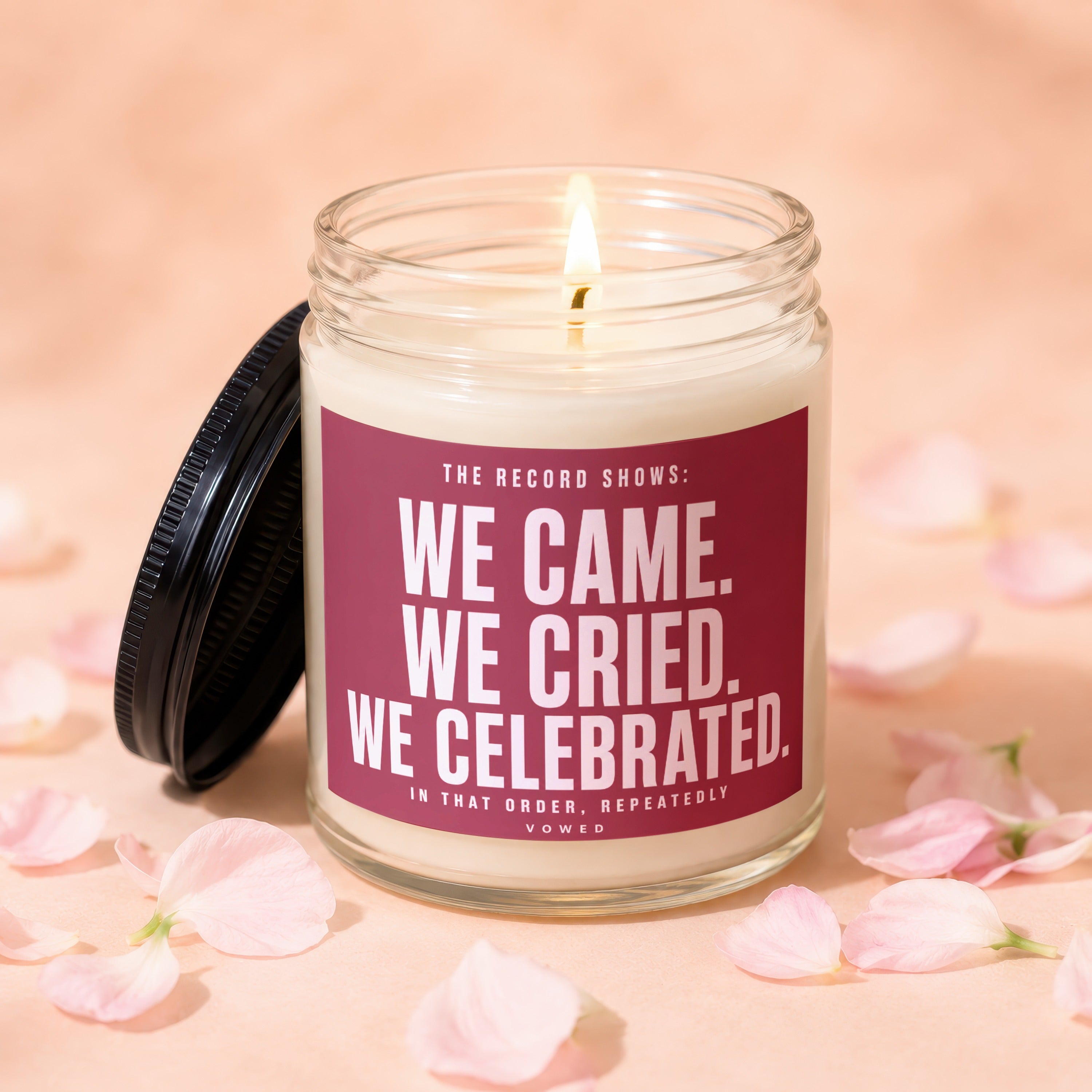Glass candle with dark lid, lit wick, and text "WE CAME. WE CRIED. WE CELEBRATED. VOWED."