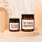 Two amber glass candles labeled "The Coven Approves" with black lids.