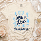 A cream-colored t-shirt with blue text reading Snow in Love Olivia's Bachelorette Park City UT, surrounded by winter and bachelorette-themed icons.
