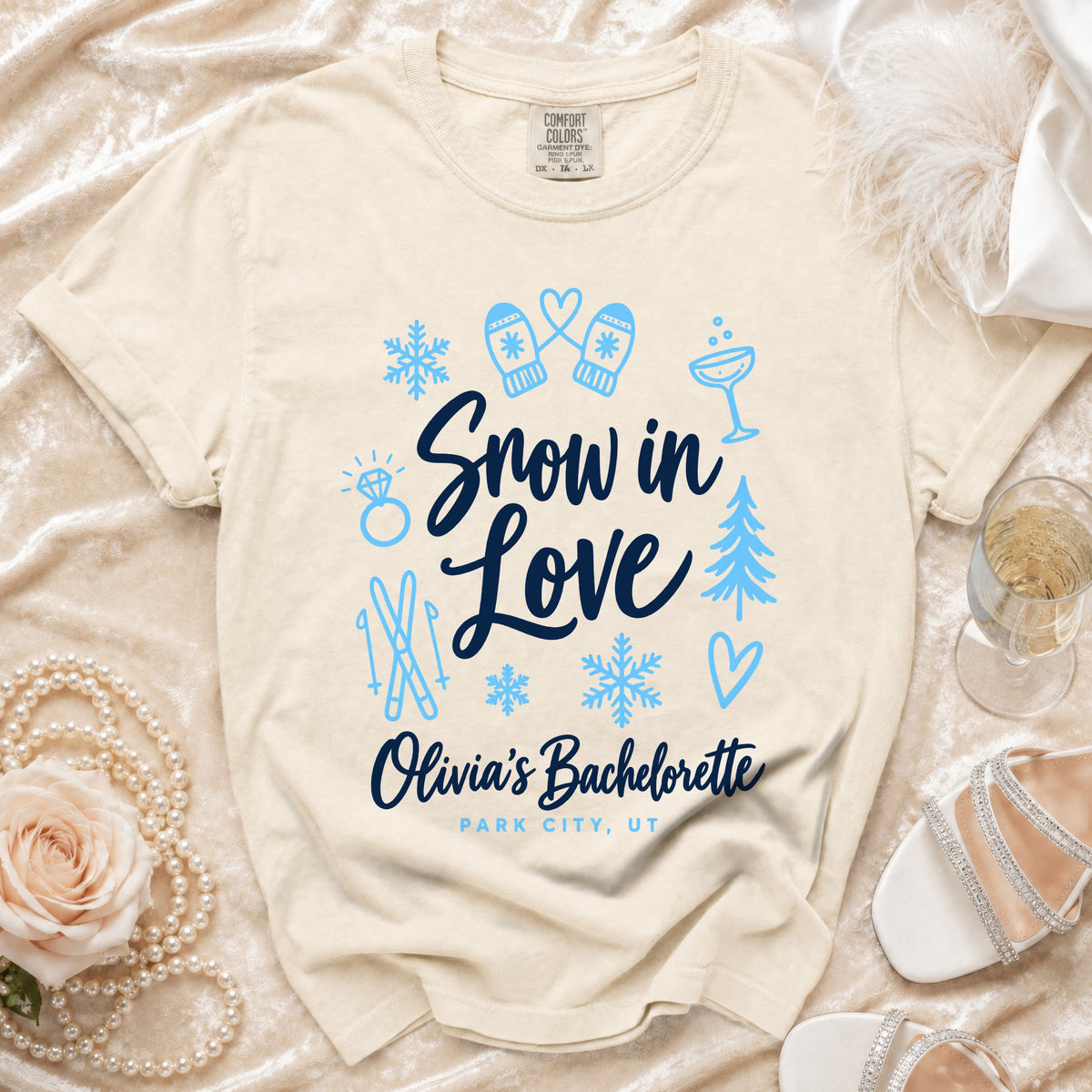 A cream-colored t-shirt with blue text reading Snow in Love Olivia's Bachelorette Park City UT, surrounded by winter and bachelorette-themed icons.