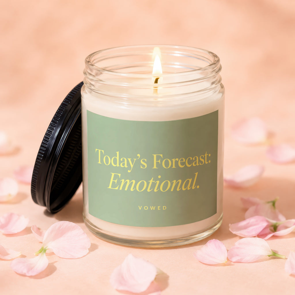 Glass candle with lit wick, featuring a label that reads "Today's Forecast: Emotional. VOWED."