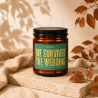 Amber jar candle with green label reading "STATUS: WE SURVIVED THE WEDDING"