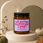Amber jar candle with black lid labeled "Will you hold my dress while I pee?"