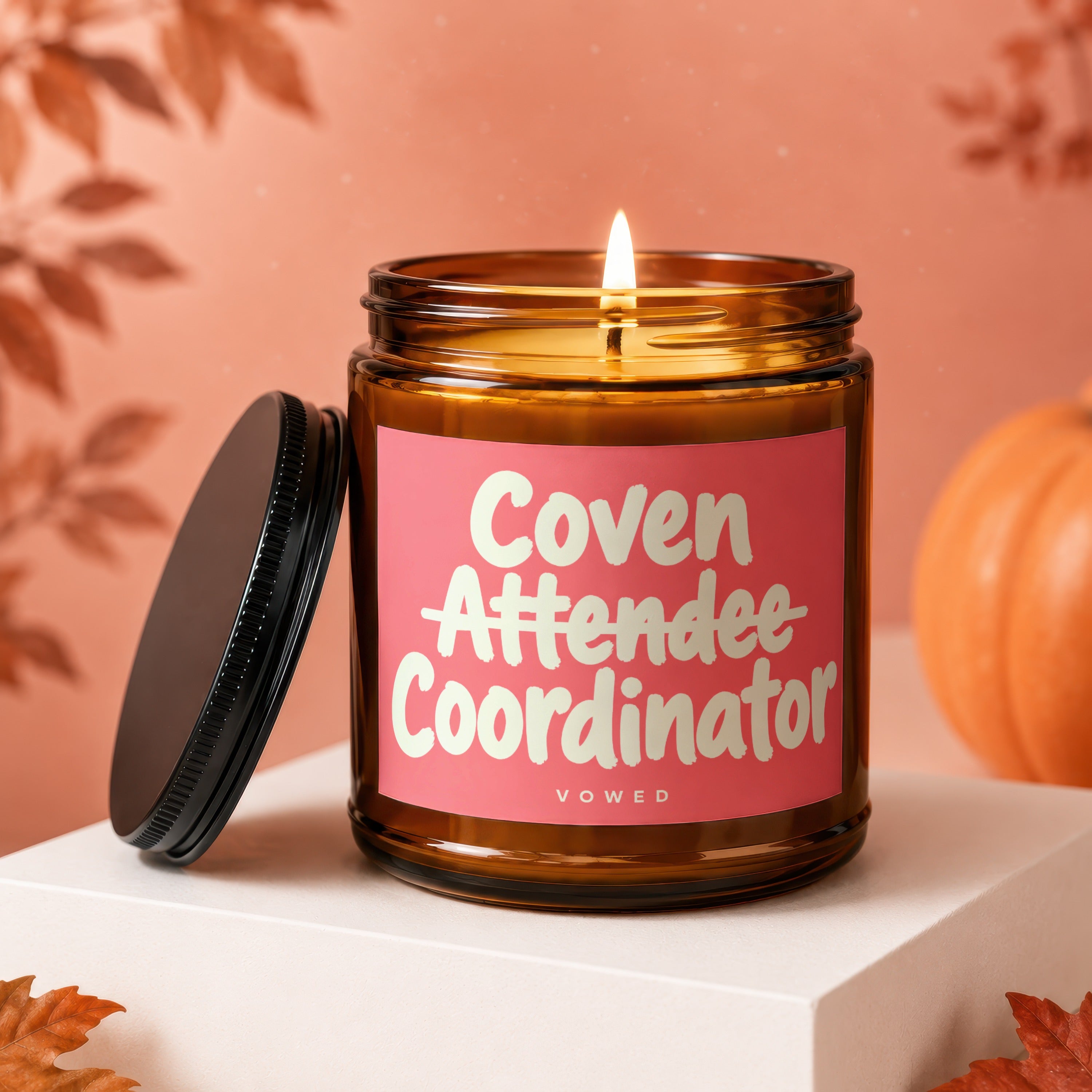 Lit amber glass candle with pink label reading "Coven Attendee Coordinator Vowed".