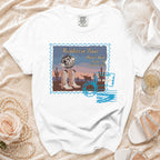 White t-shirt with "Reckless in Texas" postcard design featuring boots, disco ball, and desert scene.