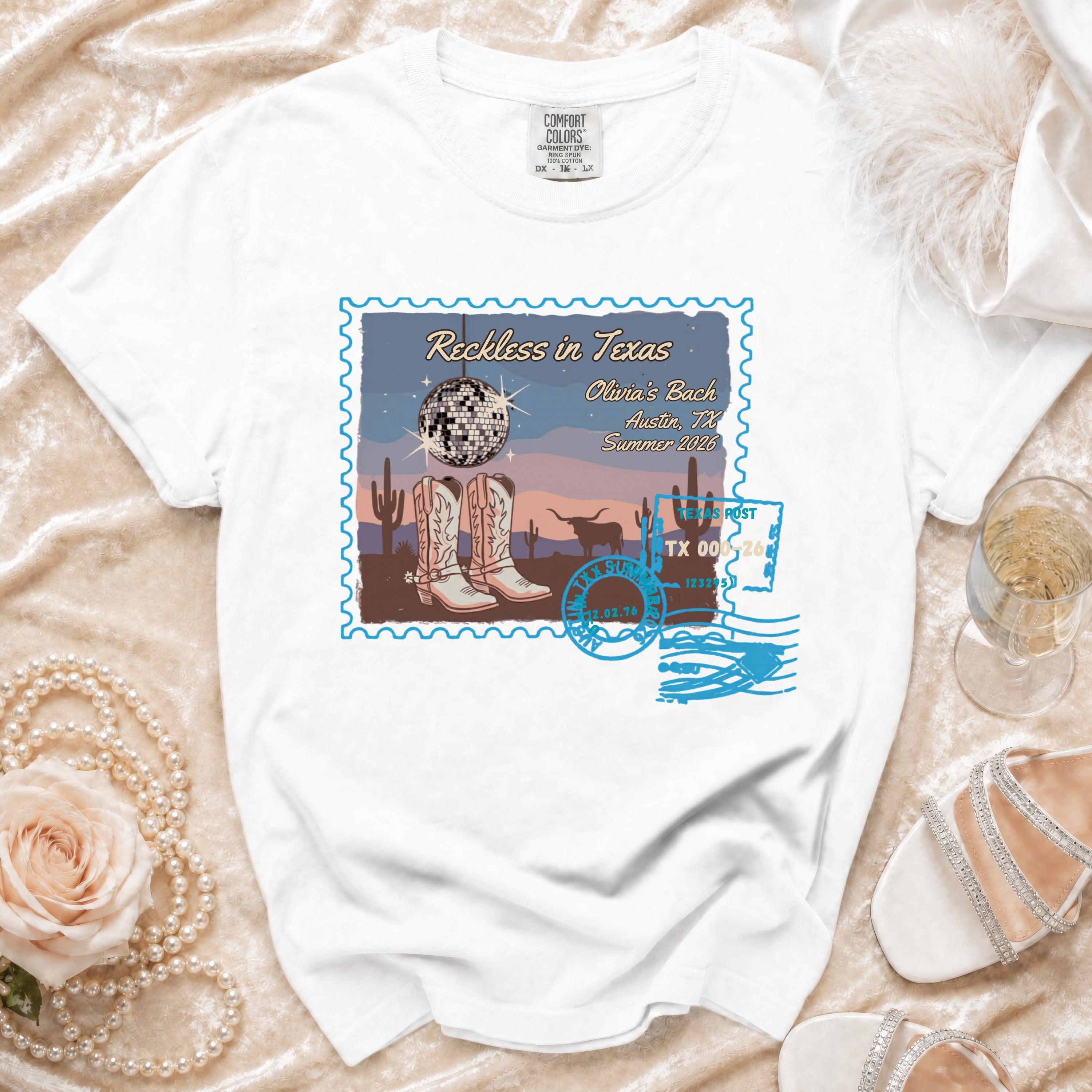 White t-shirt with "Reckless in Texas" postcard design featuring boots, disco ball, and desert scene.