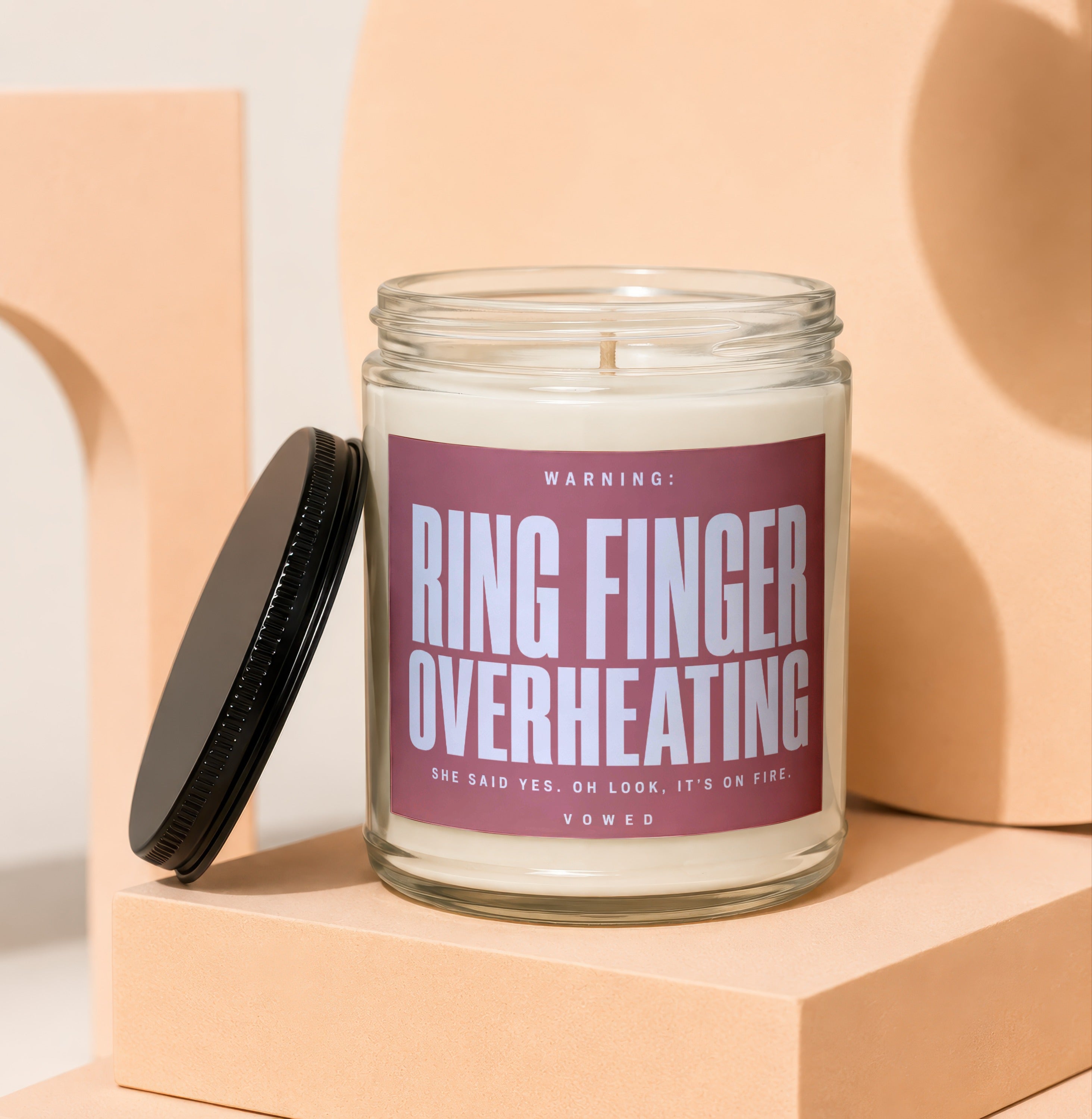 Glass candle with pink label reading "Ring Finger Overheating" and "She Said Yes. Oh Look, It's On Fire."