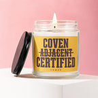 Candle in glass jar with burning wick and black lid, label reads "Coven Adjacent Certified Vowed".