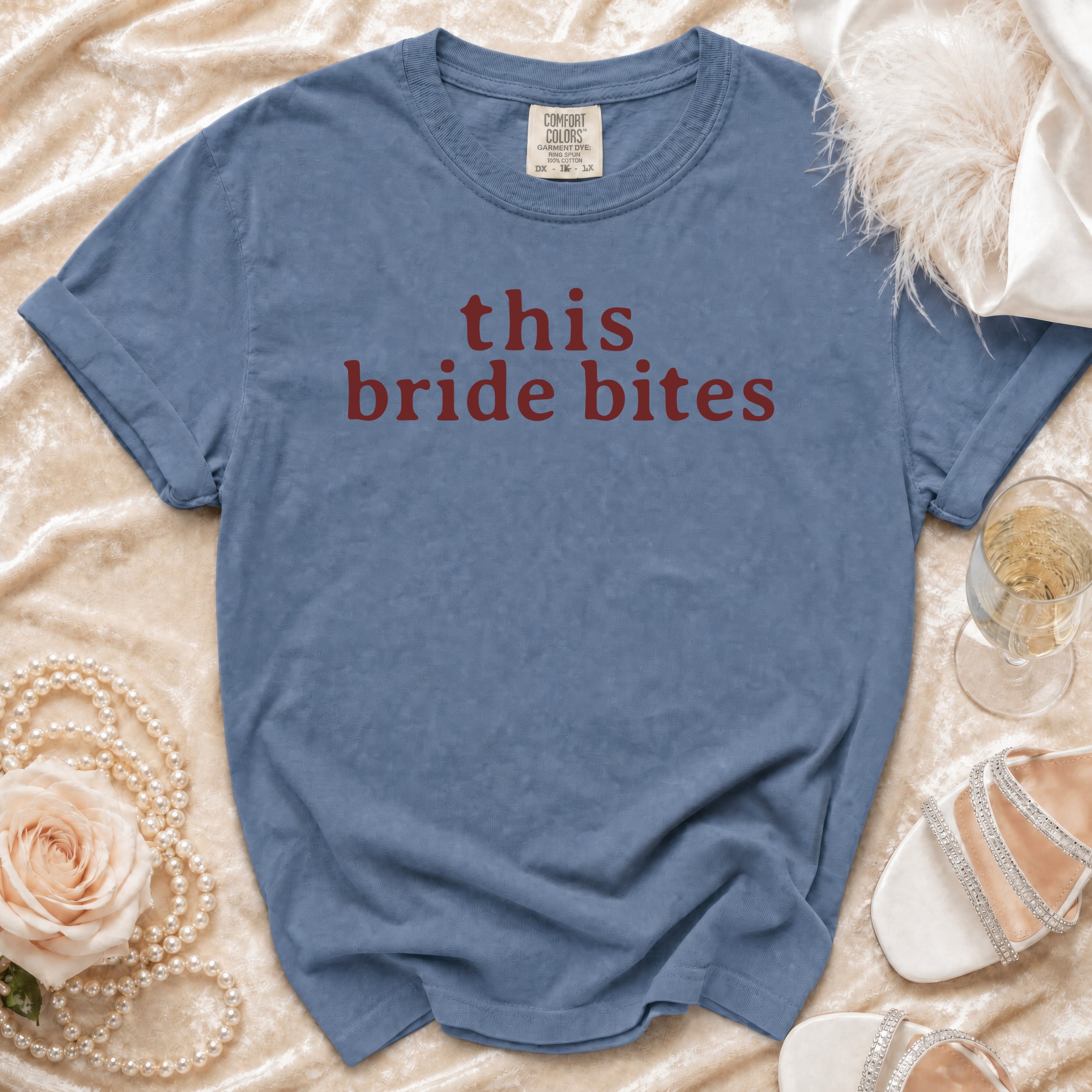 Blue t-shirt with "this bride bites" in red text, styled with pearls, a rose, and champagne.
