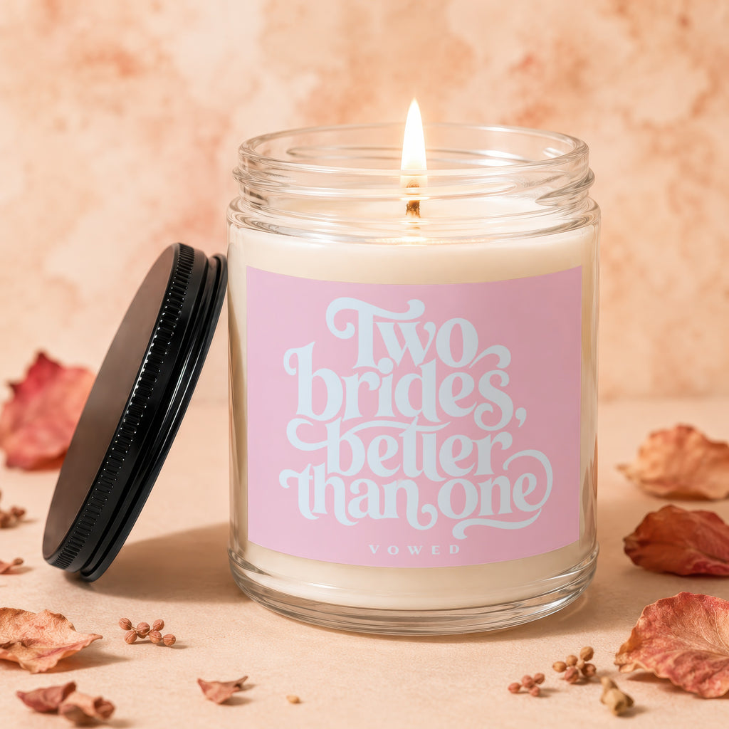 Glass candle with lit wick and pink label reading "Two brides, better than one"
