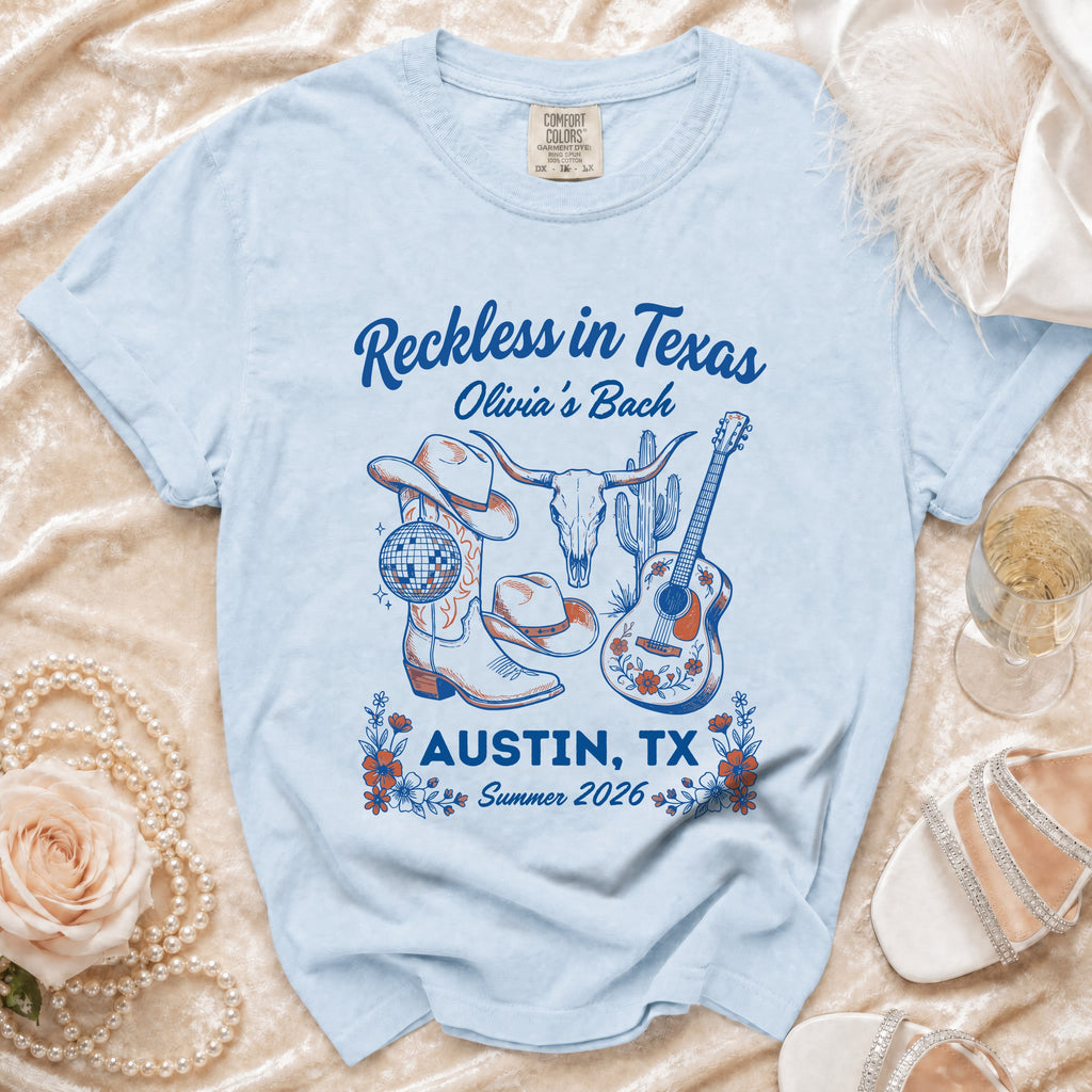 Light blue t-shirt with "Reckless in Texas Olivia's Bach Austin, TX Summer 2026" text and graphics.
