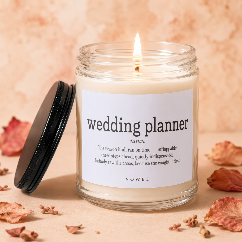 Glass candle with "wedding planner" definition label and burning wick. Flanking leaves and decor.