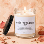 Glass candle with "wedding planner" definition label and burning wick. Flanking leaves and decor.