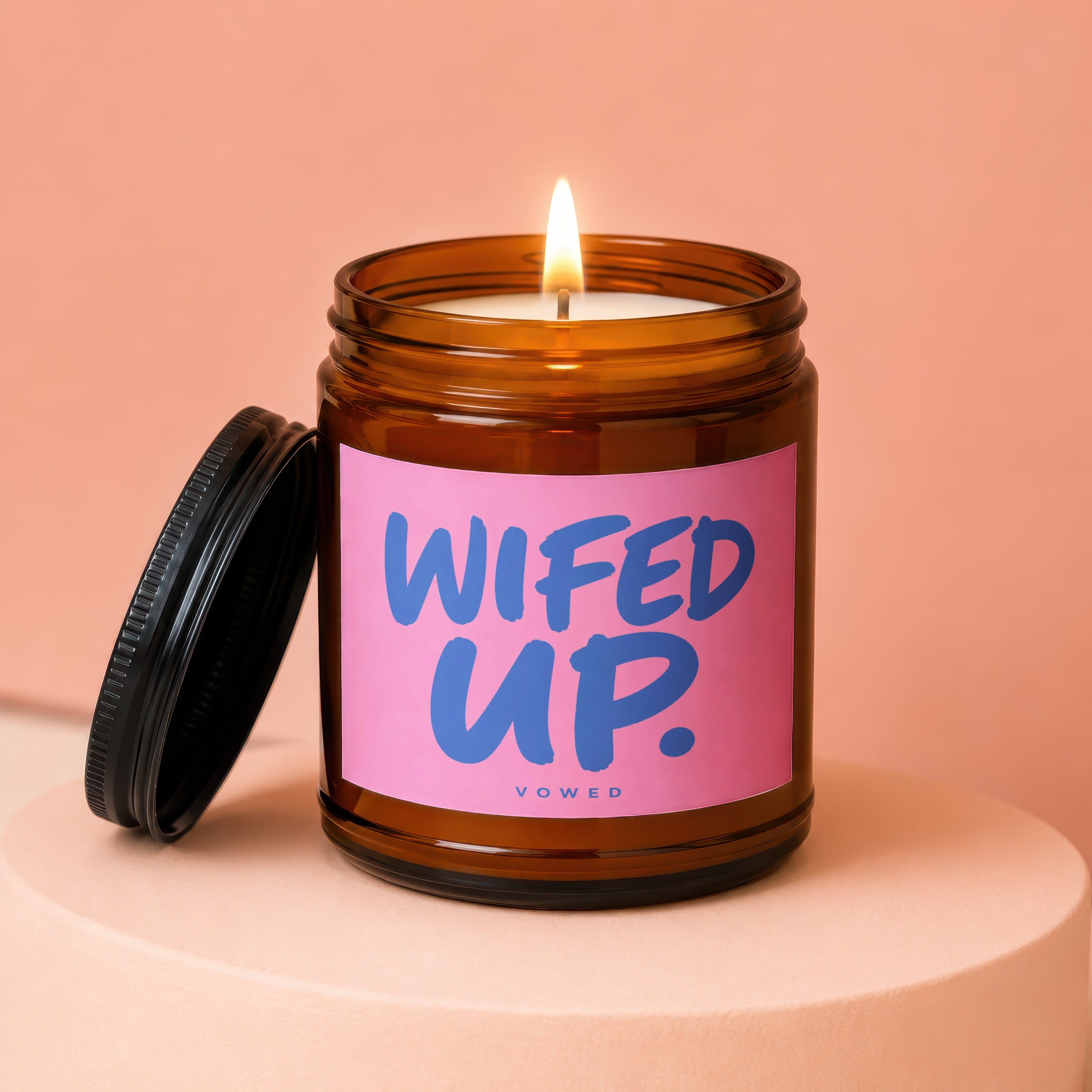 Amber jar candle with burning wick and pink label reading "WIFED UP" and "VOWED".