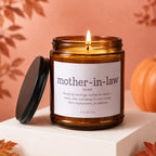 Amber candle with lit wick, "mother-in-law" definition on label, autumn leaves, and pumpkin decoration.