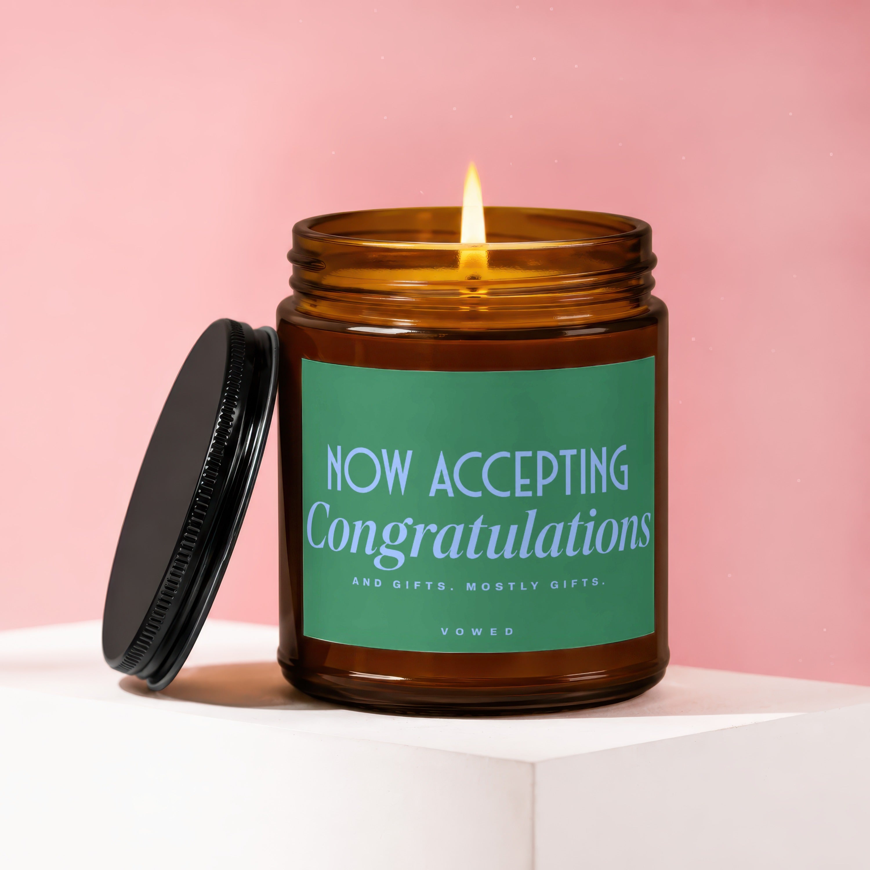 Lit candle in amber jar with green label reading "NOW ACCEPTING Congratulations"