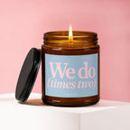 Amber jar candle with lit wick, lid to the side, reads "We do (times two)".