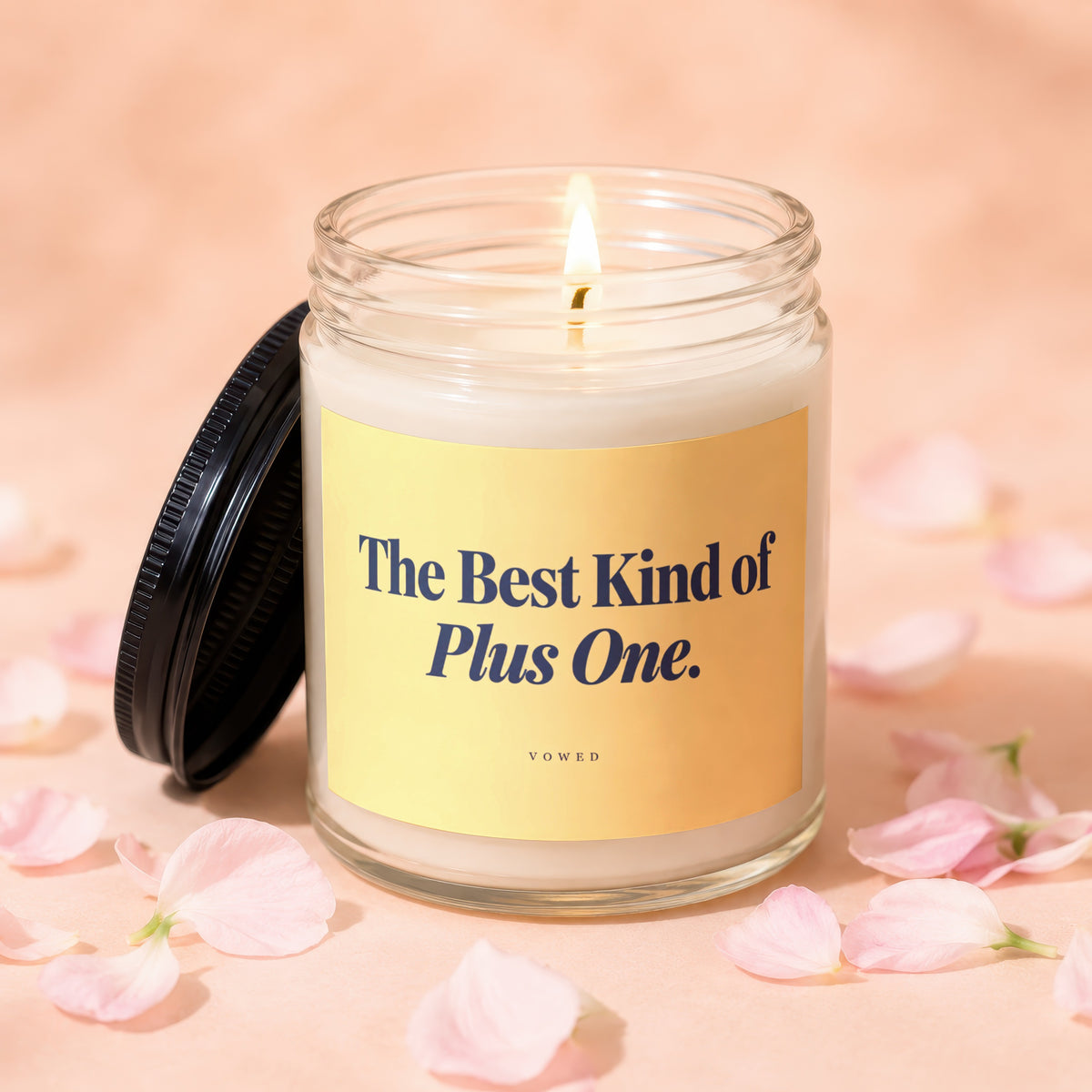 Glass candle with lit wick, yellow label reading "The Best Kind of Plus One. VOWED".