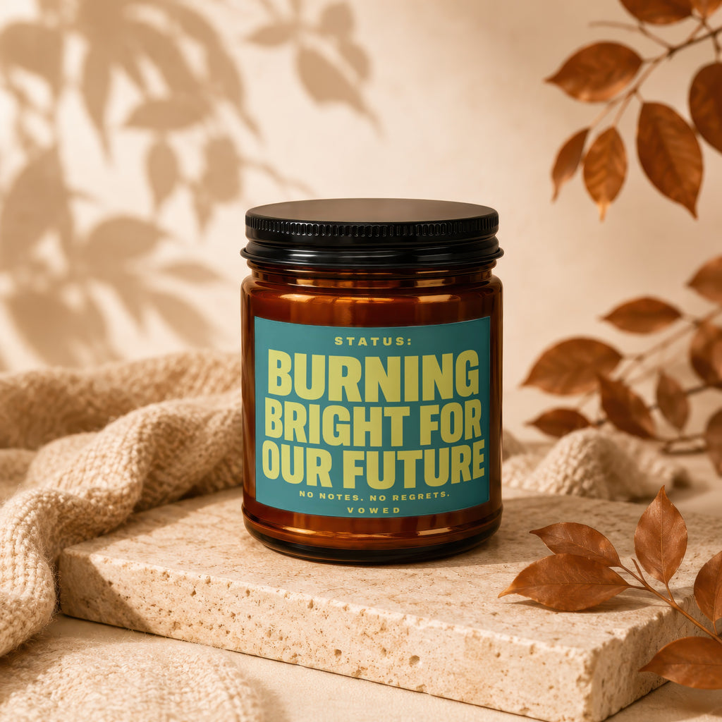 Amber jar candle with teal label reading "STATUS: BURNING BRIGHT FOR OUR FUTURE".
