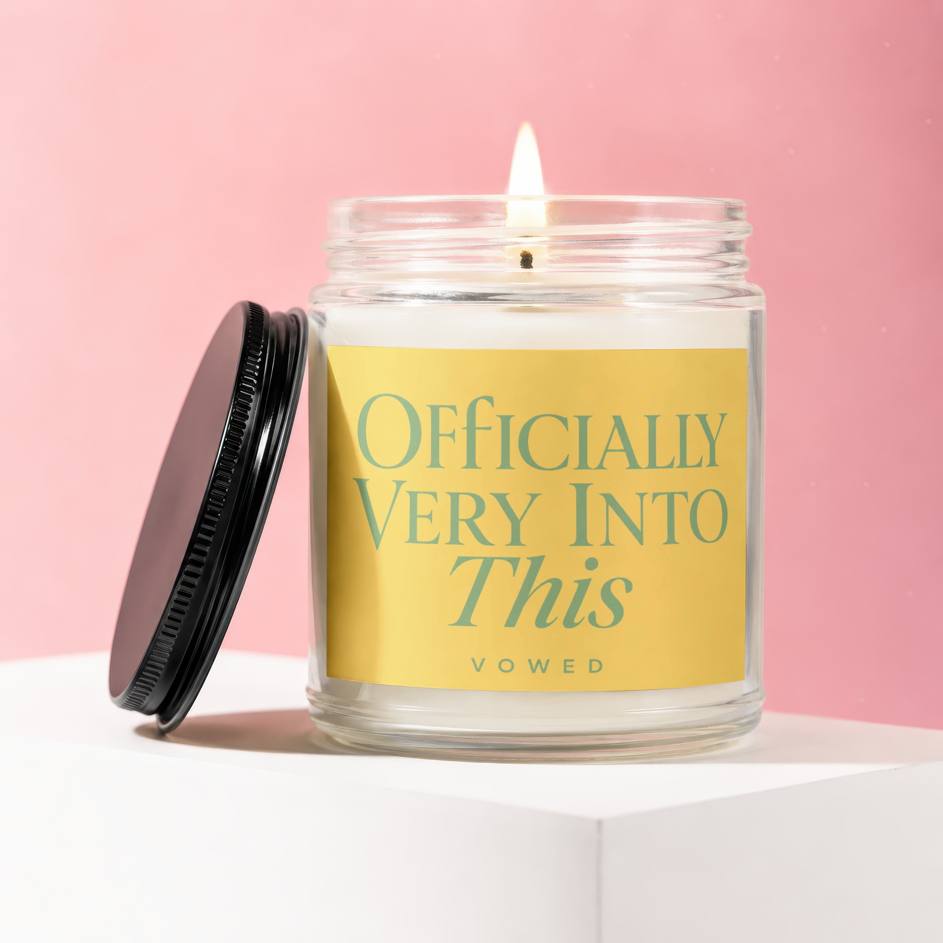 Glass jar candle with lit wick and "Officially Very Into This vowed" text on yellow label.