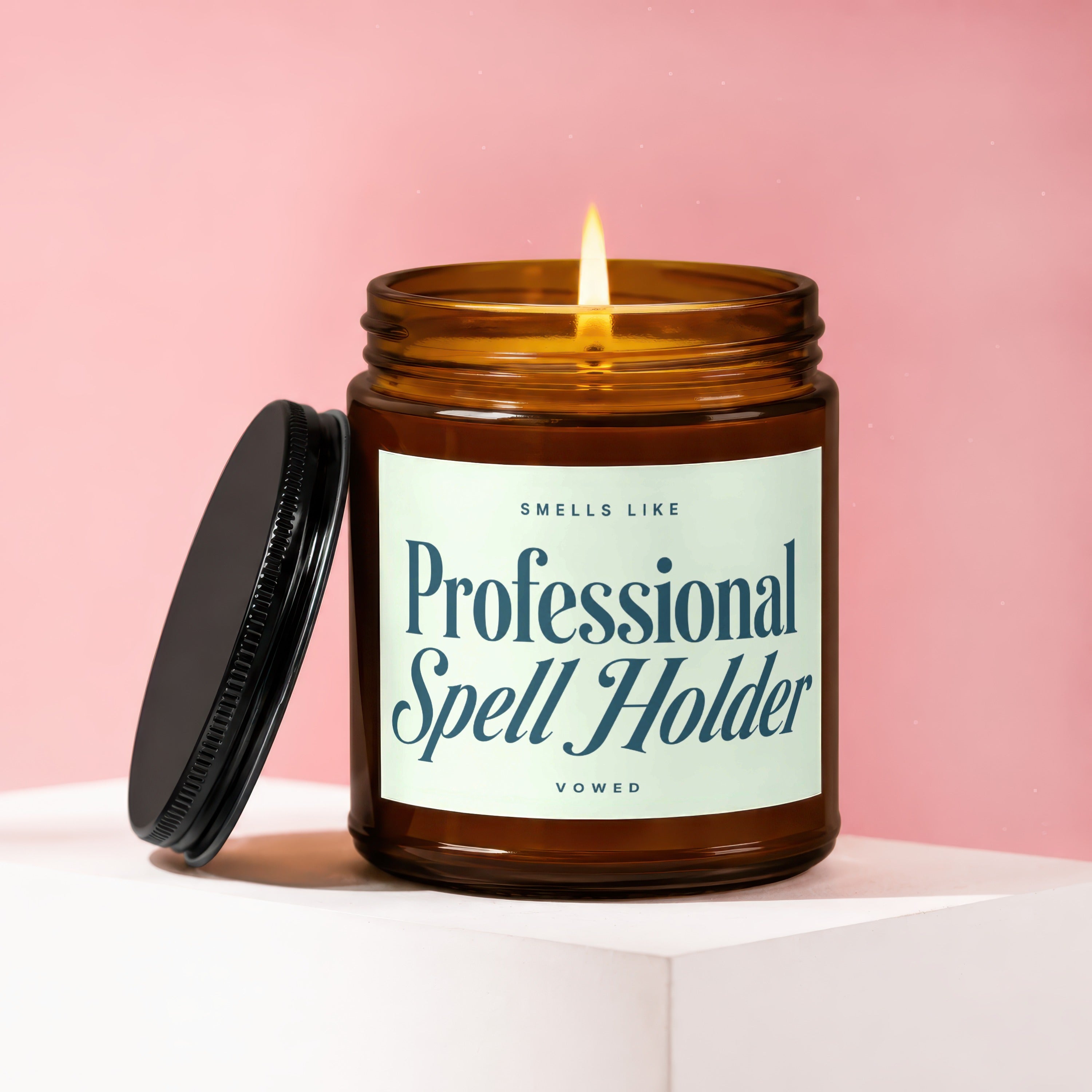 Amber glass candle with lit wick, black lid, and label reading "Smells Like Professional Spell Holder Vowed".