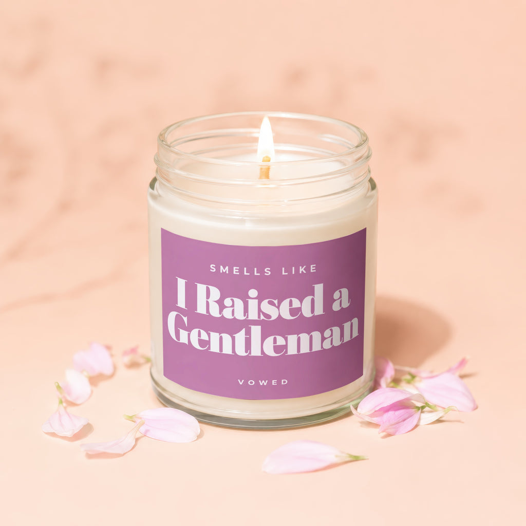 Lit candle with "I Raised a Gentleman" text on a purple label, surrounded by petals.