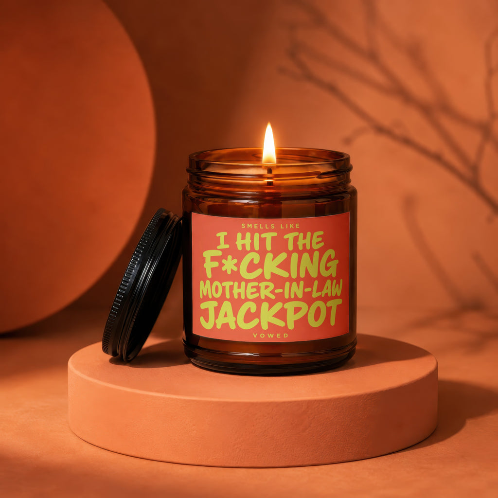 Amber jar candle with lit flame, reads "Smells like I hit the f*cking mother-in-law jackpot".