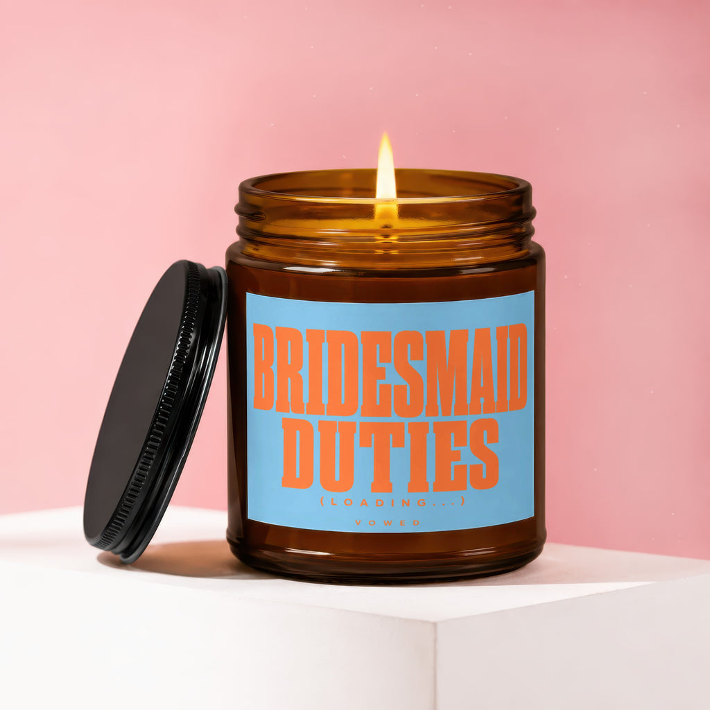 Amber jar candle with lit wick, lid nearby, reads "BRIDESMAID DUTIES (LOADING...)" and "VOWED".