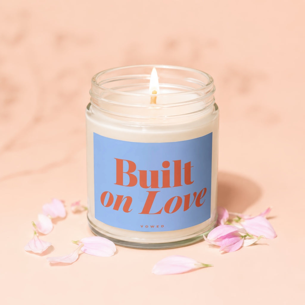 Lit candle in jar with "Built on Love" label surrounded by pink petals.
