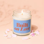 Lit candle in jar with "Built on Love" label surrounded by pink petals.