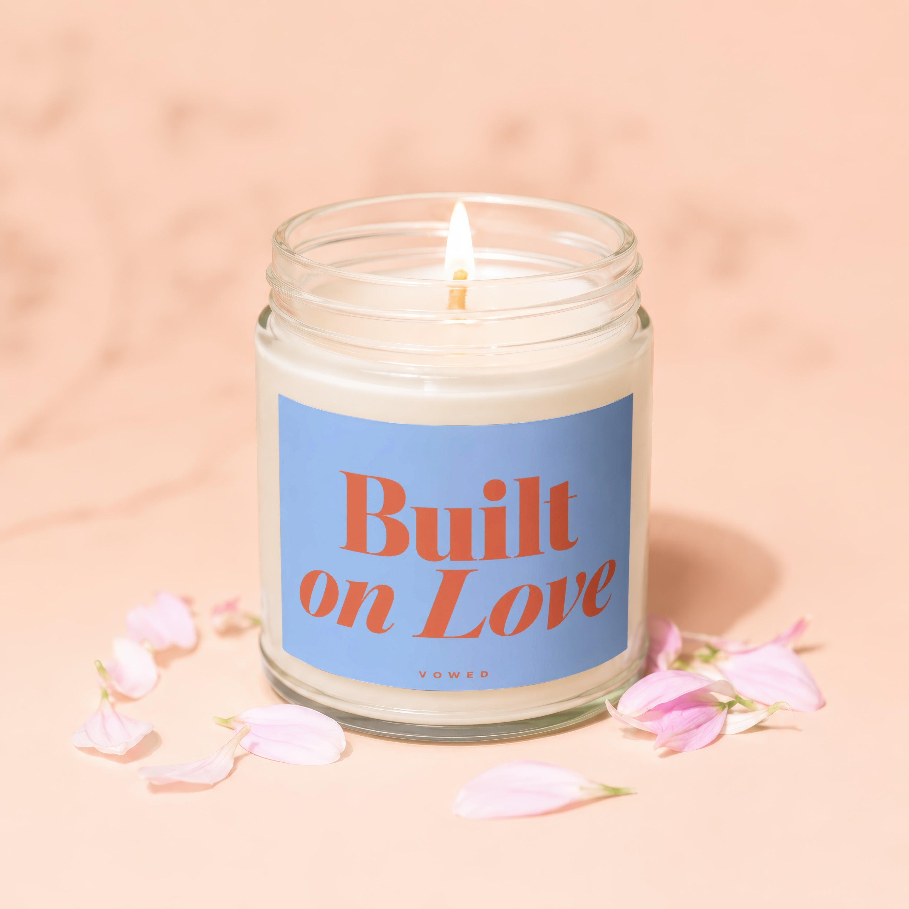 Lit candle in jar with "Built on Love" label surrounded by pink petals.