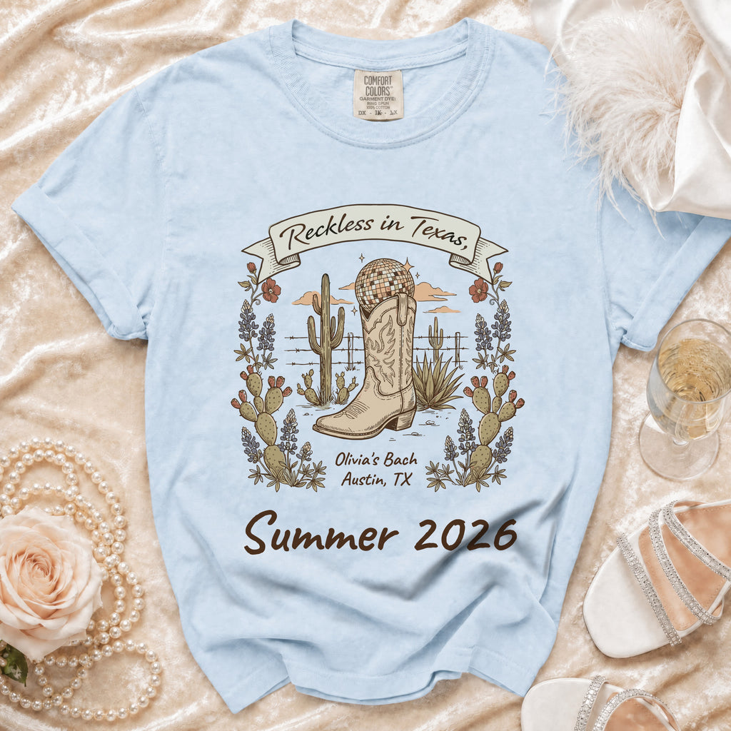 Light blue t-shirt with cowboy boot, cactus, and text "Reckless in Texas, Olivia's Bach Summer 2026".