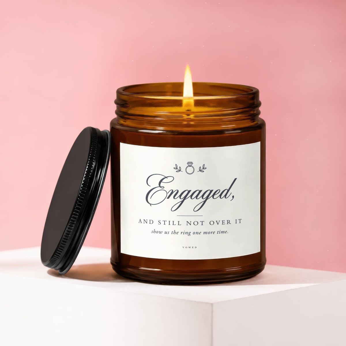 Amber glass candle with lit wick, black lid, and label that says "Engaged, AND STILL NOT OVER IT".