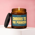 Amber glass candle with lit flame, lid ajar, reads "Cheers to the Fiancés".